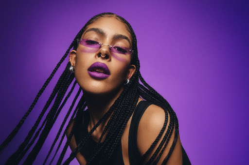 Headshot of a stylish adult female with box braids, purple lipstick, and sunglasses against a purple gradient background.