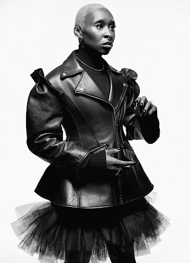 Black and white portrait of a short-haired woman wearing a black leather jacket with a tulle skirt, posing in a photo studio.