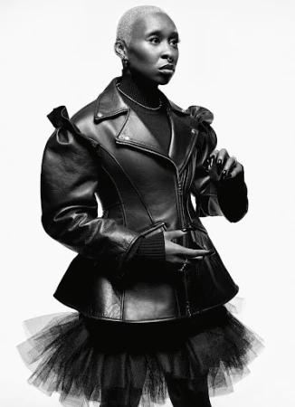 Black and white portrait of a short-haired woman wearing a black leather jacket with a tulle skirt, posing in a photo studio.