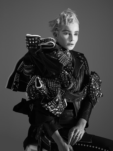 Monochrome portrait of a person with a platinum blonde hairstyle wearing a studded leather jacket and posing in a photo studio.