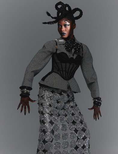 Adult female fashion model with sculptural hairstyle, dark eye makeup, statement jewelry, and patterned skirt and jacket posing in studio.