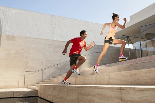 Two runners, one male in red and one female in pink, ascend outdoor stairs in athletic wear, under bright sunlight and clear blue sky.