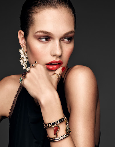 Portrait of a young adult woman with dark hair wearing ornate jewelry, including earrings, rings, and bracelets, against a gray background.