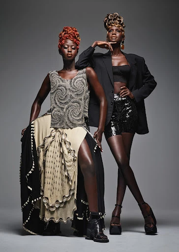 Two dark-skinned fashion models pose in a studio setting wearing stylish clothing, with one seated and the other standing.