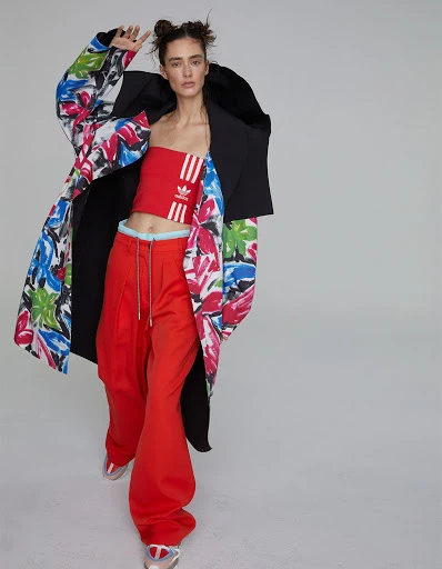 Young woman wearing a red Adidas top and red pants with a floral coat posing in a studio setting with a neutral background.