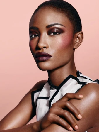 Close-up beauty shot of a young, dark-skinned Black woman with dramatic makeup, looking at the camera against a soft peach background.