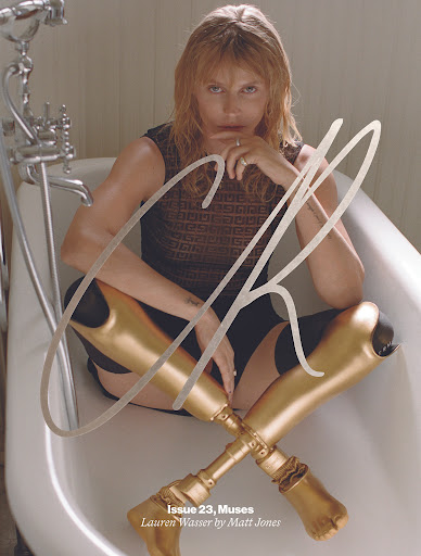 Adult woman with prosthetic legs poses in a bathtub for magazine cover. She wears a dark top and sits confidently, looking directly at the camera.