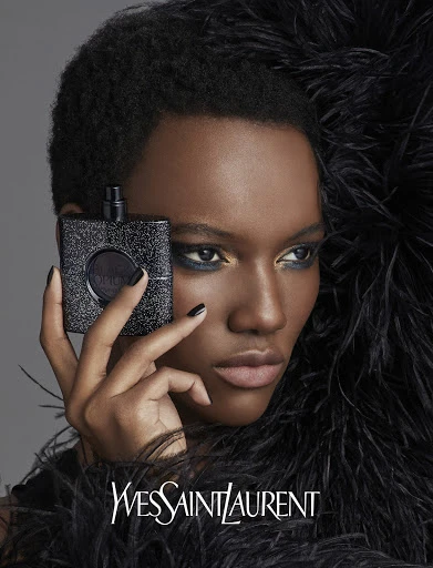 Close-up shot of a Black woman holding a black square perfume bottle, wearing a feathered top, with the brand Yves Saint Laurent visible.