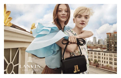 Two fair-skinned women pose on a rooftop in an outdoor fashion shot, with a black handbag and city skyline backdrop.