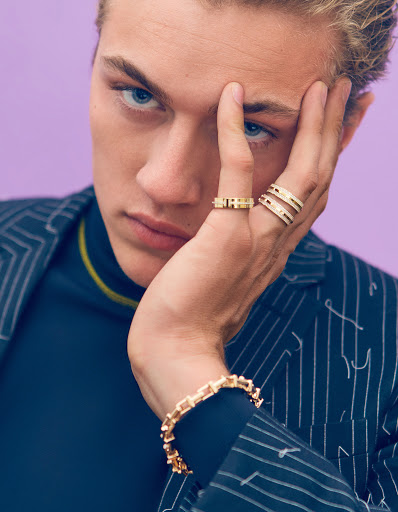 Close-up portrait of a blond male model wearing a navy blazer and jewelry including a bracelet and rings against a purple background.