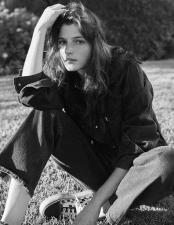Black and white portrait of a woman seated on grass wearing dark denim and sneakers, styled by Daniel Edley