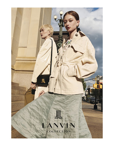 Two fashion models wearing coats and skirts are posing on a city sidewalk in front of a building with a Lanvin Collection logo.