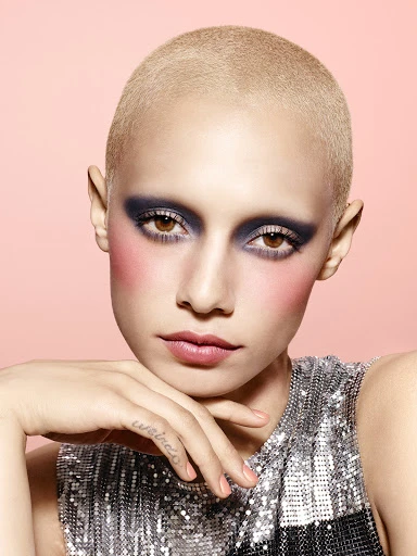 Close-up portrait of a bald woman with dramatic dark eyeshadow and blush, wearing a reflective top, against a light pink background.