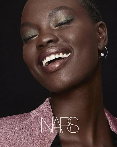 Close-up portrait of a smiling African-American woman with green eyeshadow wearing a sparkling pink blazer, against a dark background.