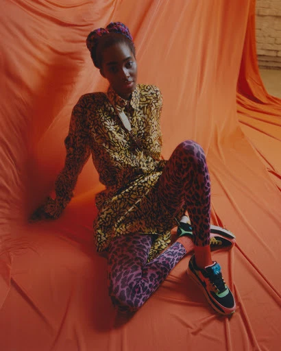 Young woman with dark skin poses in a patterned shirt and leggings on an orange textile backdrop with black, green, and white sneakers.