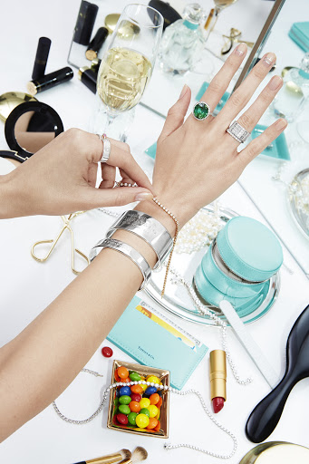 Woman's hands putting on a bracelet surrounded by makeup, jewelry, and candy on a white surface, creating a luxurious, styled shot.
