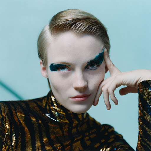 Close-up portrait of a blonde adult female model with dark eye makeup wearing a tiger-striped sequin top against a light blue background.