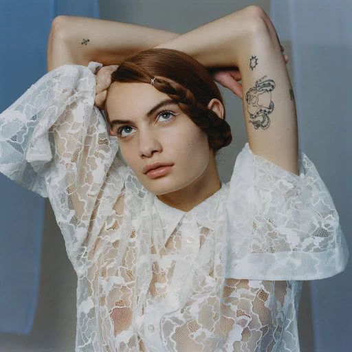 Portrait of a young adult female with braided hair, wearing a white lace shirt and posing in front of a blue and white background.