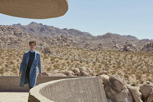 An adult male model in a blue coat stands on a structure in a desert landscape with mountains under a clear blue sky on a sunny day.