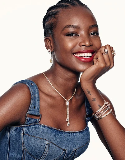 Smiling dark-skinned woman with braided hair poses in a denim dress with diamond jewelry against a white backdrop, medium shot.