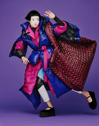 Fashion editorial shot of a young woman with exaggerated makeup wearing a blue and pink puffer coat and carrying a quilted tote.