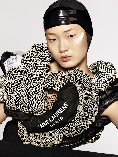 Close-up of a model wearing an avant-garde black head covering and sculptural mesh collar. The photograph features a white studio backdrop.