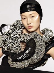 Close-up portrait of an East Asian model wearing a black swim cap and sculptural textured white and black braided apparel with Saint Laurent branding.