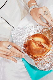 Close-up of hands wearing silver rings and bangles unwrap a bagel on foil styled by Daniel Edley