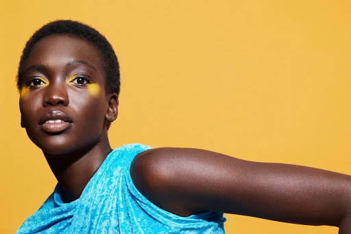 Close-up portrait of a Black woman with short dark hair wearing a bright blue top and yellow eye makeup against a solid yellow background
