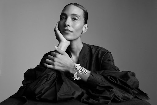 Black and white portrait of a woman with slicked-back hair wearing butterfly-shaped jewelry, a dark top, and neutral makeup in studio lighting.