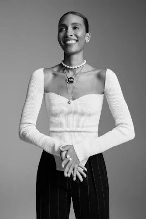Black woman smiling wearing a ribbed white off-shoulder top and layered necklaces against a solid grey background