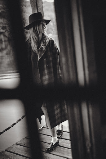 Black and white portrait of a woman in a hat and coat walking on a wooden pier with blurred foreground elements adding depth.