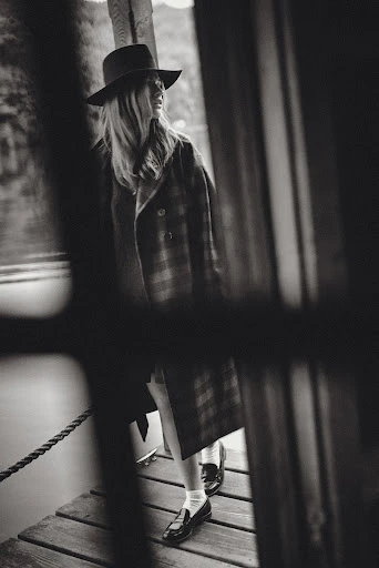 Black and white portrait of a woman in a hat and coat walking on a wooden pier with blurred foreground elements adding depth.