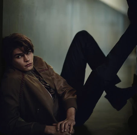 Portrait of a young adult male model in a brown jacket and dark pants, reclining against a wall indoors with soft, diffused lighting.