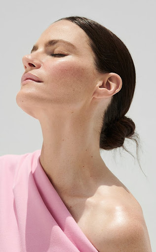 Close-up profile portrait of a woman with her eyes closed, bathed in sunlight wearing a pink garment against a light background.