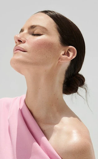 Close-up profile portrait of a woman with her eyes closed, bathed in sunlight wearing a pink garment against a light background.