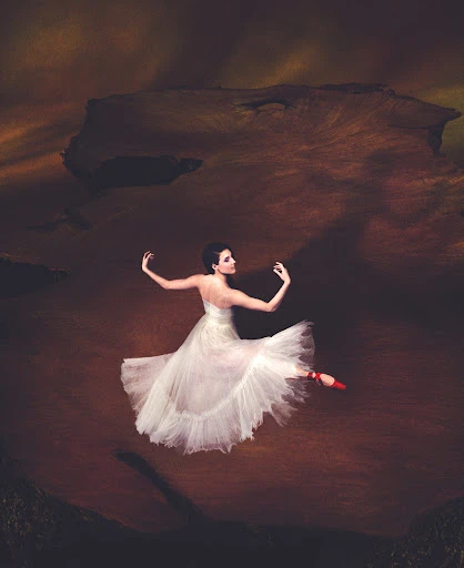 Elegant ballerina in white tutu and red ballet slippers performs ballet mid-air in a dramatic indoor setting with dark, moody lighting.