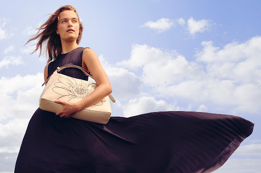A woman in a flowing dark dress holds a tan purse with a flower design, standing against a cloudy sky in an outdoor fashion shot.