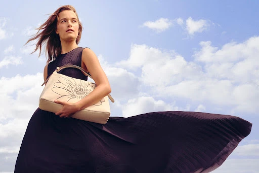 A woman in a flowing dark dress holds a tan purse with a flower design, standing against a cloudy sky in an outdoor fashion shot.