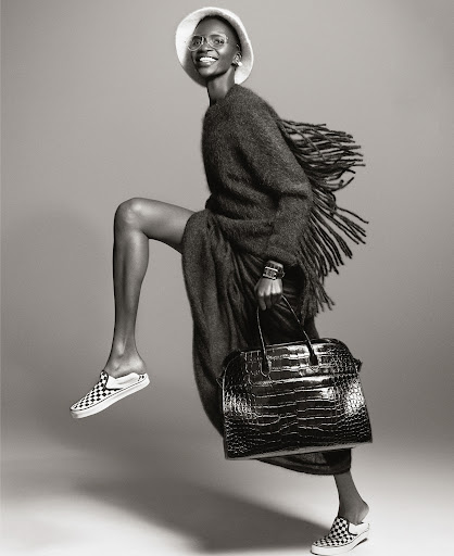A fashion shot in black and white featuring an African-American woman in motion, wearing a hat, oversized cardigan and carrying a handbag.