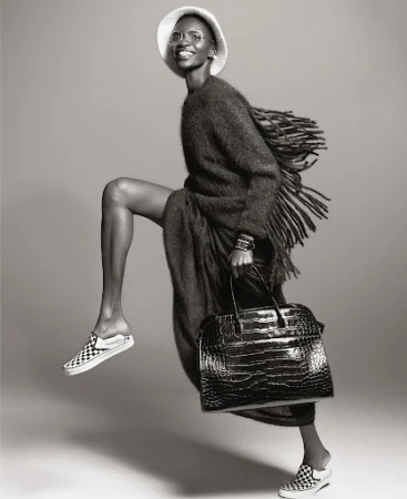 A fashion shot in black and white featuring an African-American woman in motion, wearing a hat, oversized cardigan and carrying a handbag.