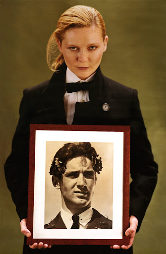 Woman in black suit holds framed vintage portrait. Olive green background, formal wear, portrait photography, classic style, fair skin.