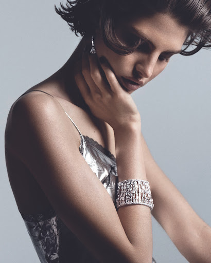 Portrait of a brunette woman with hand near face, wearing a shiny silver dress and statement jewelry in a photo studio setting.