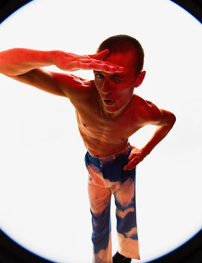 Low angle shot of a shirtless adult man with blue dyed pants, winking and holding hand above eyes as he poses in photo studio.