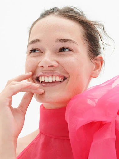 Smiling woman in a pink top with bow detail looking away in a studio portrait, showcasing fashion and beauty with a focus on light.