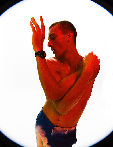 Red-toned portrait of a bare-chested man with a wristwatch in an expressive pose against a bright white backdrop.