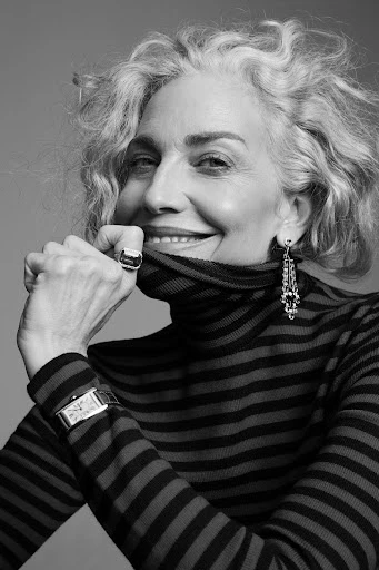 Black and white portrait of a smiling mature woman with curly gray hair wearing a striped turtleneck sweater, large ring, and dangling earrings.
