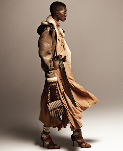 A fashion shot captures a Black woman in a brown coat and dress with a zebra-striped purse, set against a neutral backdrop in a studio.