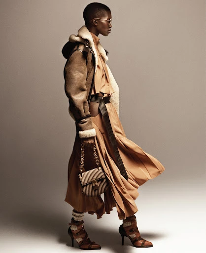 A fashion shot captures a Black woman in a brown coat and dress with a zebra-striped purse, set against a neutral backdrop in a studio.