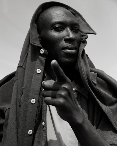 Monochromatic portrait of a Black adult male in a hooded jacket pointing directly at the camera, set against a plain, bright sky.